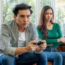 Man and woman sitting on couch with man holding a PlayStation controller and his partner looking at him disapprovingly