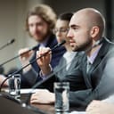 One of young speakers talking in microphone at business or political conference between colleagues