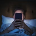 Anonymous Man Uses Smartphone in Bed at Home at Night. Handsome Guy Browsing Social Media, Reading News, Doing Online Shopping Late at Night. Focus on Hand Holding Mobile Phone Covering Face