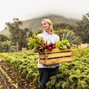 young female chef smiling while holding a crate of fresh vegetables while standing in an organic farm field