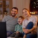 family, leisure and people concept - happy smiling father, mother and little son with remote control watching tv at home at night