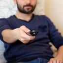 remote control in focus as a young man sits on a couch in his living room, browsing TV channels