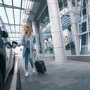 Beautiful young woman standing with a trolley suitcase next to the car at airport terminal.
