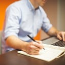 Man writing down notes as he works on laptop