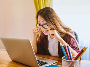A young woman looking at a laptop while biting a pen