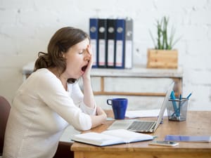 A young woman at an office desk holding her hand over her eyes and yawning