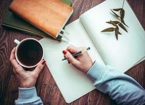 a woman writing in a notebook with her right hand and holding a cup of coffee in her left