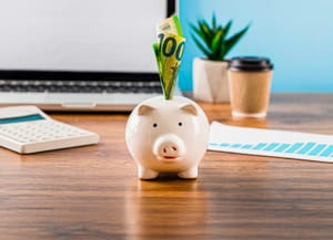 Piggy bank with a $100 bill on top of it placed on a table next a calculator and laptop
