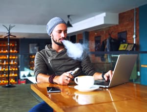 A young man vaping in a cafe while looking at his laptop