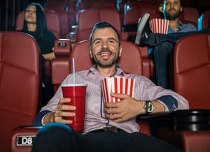 A guy enjoying himself at the movies, holding a large soda in one hand and popcorn in the other.