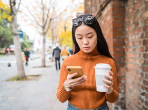 A young, teenage girl looking at her phone and holding a cup of coffee.