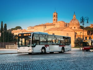 A public bus cruises along Via dei Fori Imperiali on a sunny summer morning in Rome, Italy.