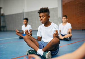 An African American schoolboy and his friends meditating in lotus position during a PE class at school gym.