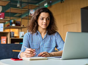 A Hispanic college student taking down notes and looking at her laptop.