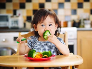 a toddler wearing a chef's hat and apron sitting at a kitchen table, grimacing after tasting something