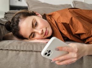 A young woman lying on the sofa and looking at her smartphone with a somewhat bored expression on her face.