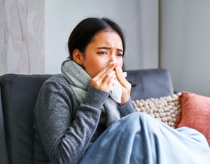 A sick woman sitting at home, covered in warm clothes and a scarf, blowing her nose 