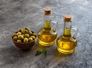Two glass bottles of olive oil with cork stoppers, placed on a textured gray surface.