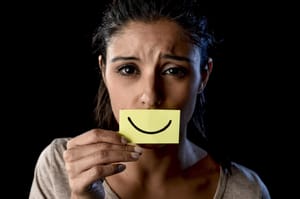 Depressed girl hiding her mouth behind a piece of paper with a smile drawn on it.