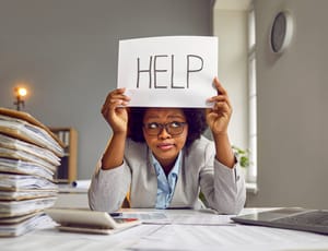A tired and busy businesswoman in a suit and glasses sitting at an office desk with a load of paperwork and holding a paper sign with the word HELP.