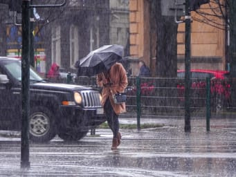 A woman in a brown coat holding an umbrella crosses a busy street in heavy rain, amid heavy precipitation and flooding of city streets.