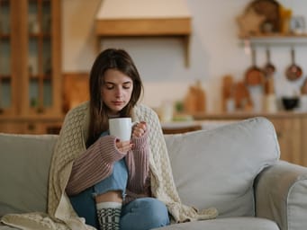 A young woman wearing a knitted sweater, sitting on the sofa at home, wrapped in a plaid blanket, drinking hot coffee or tea. 