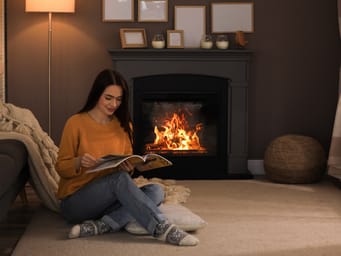 A beautiful young woman reading a magazine on the floor near a cozy fireplace at home.
