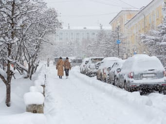 A snow-covered city street during a heavy snowfall in Russian Far East, with snow on the sidewalk, cars, and tree branches, as women walk around the winter city in snowy weather.