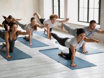 A young attractive Indian woman leads a yoga class in a spacious, light-filled room with a grey color scheme. 