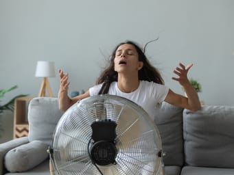 A young Caucasian woman sitting on a couch in the living room, breathing fresh air from a floor fan, struggling with the heat due to the lack of air conditioning during summer weather.